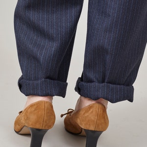K&ouml;nnte beinhalten: Ein Paar braune Wildlederschuhe mit Absatz und einer Schleife vorne. Die Schuhe werden zu einer dunkelblauen Hose mit feinen Nadelstreifen getragen.