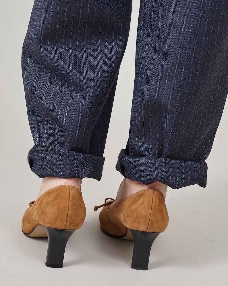 K&ouml;nnte beinhalten: Ein Paar braune Wildlederschuhe mit Absatz und einer Schleife vorne. Die Schuhe werden zu einer dunkelblauen Hose mit feinen Nadelstreifen getragen.