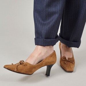 K&ouml;nnte beinhalten: Ein Paar braune Wildlederschuhe mit Schleife an der Vorderseite. Die Schuhe haben eine spitze Zehenpartie und einen niedrigen Absatz.