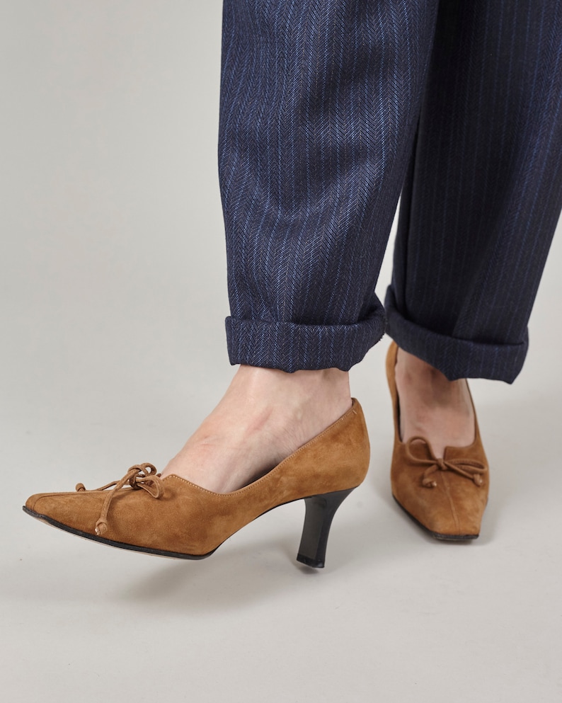 K&ouml;nnte beinhalten: Ein Paar braune Wildlederschuhe mit Schleife an der Vorderseite. Die Schuhe haben eine spitze Zehenpartie und einen niedrigen Absatz.