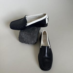 May include: Black leather loafers with white trim and a rounded toe. The shoes have a classic design with a stitched sole and a small heel. One shoe is resting on a dark gray rock, while the other is on a white surface.