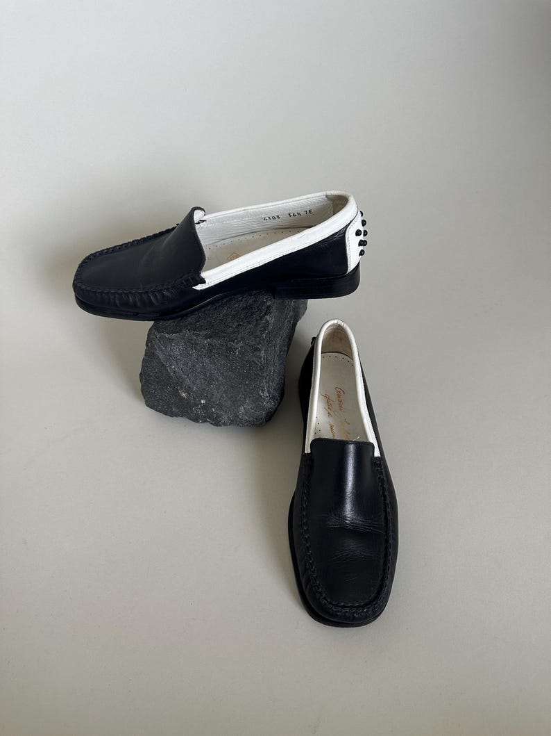 May include: Black leather loafers with white trim and a rounded toe. The shoes have a classic design with a stitched sole and a small heel. One shoe is resting on a dark gray rock, while the other is on a white surface.