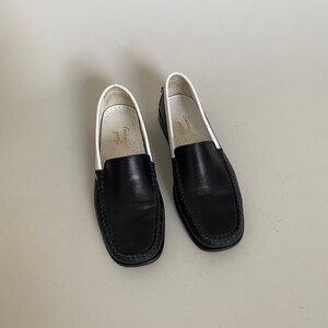 May include: A pair of black leather loafers with white trim and a stitched design around the toe. The shoes have a classic, slip-on style, suitable for various occasions. The interior of the shoe is white with gold text.