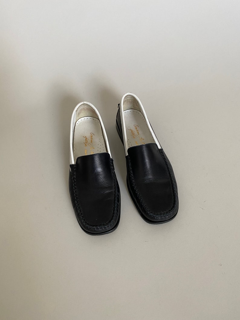 May include: A pair of black leather loafers with white trim and a stitched design around the toe. The shoes have a classic, slip-on style, suitable for various occasions. The interior of the shoe is white with gold text.