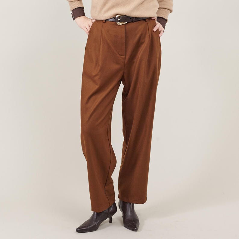 Woman wearing high-waisted chestnut brown wool trousers with pleats, a tapered cropped cut, and a relaxed yet tailored fit. Styled with a dark brown belt, a beige sweater, and dark brown pointed-toe leather ankle boots, these elegant trousers offer a sophisticated yet effortless look.