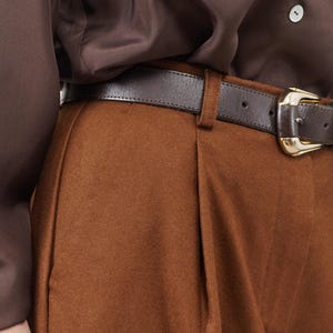 May include: Brown pants with a brown leather belt and a gold buckle. The pants have pleats in the front.