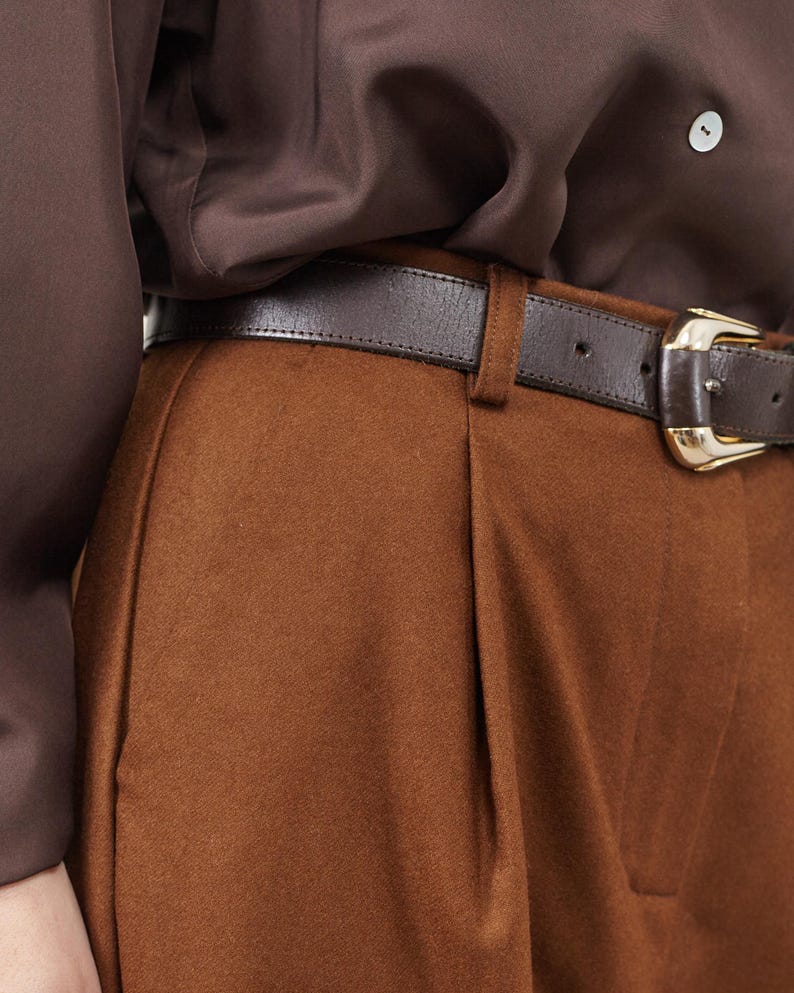 May include: Brown pants with a brown leather belt and a gold buckle. The pants have pleats in the front.