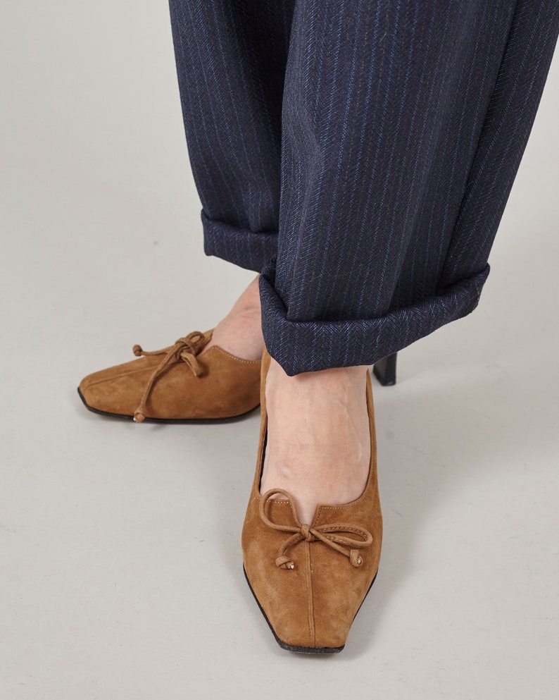 K&ouml;nnte beinhalten: Braune Wildlederschuhe mit Absatz und einer Schleife an der Vorderseite. Die Schuhe werden mit einer dunkelblauen Hose mit Nadelstreifen getragen.