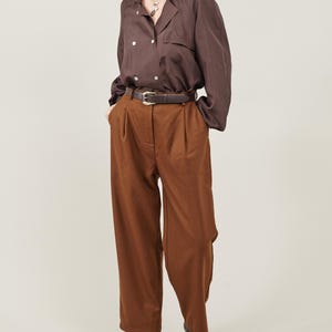 May include: A brown button-down shirt with a long sleeve and a brown leather belt. The shirt is tucked into brown wide-leg pants. The person is wearing black heeled boots.