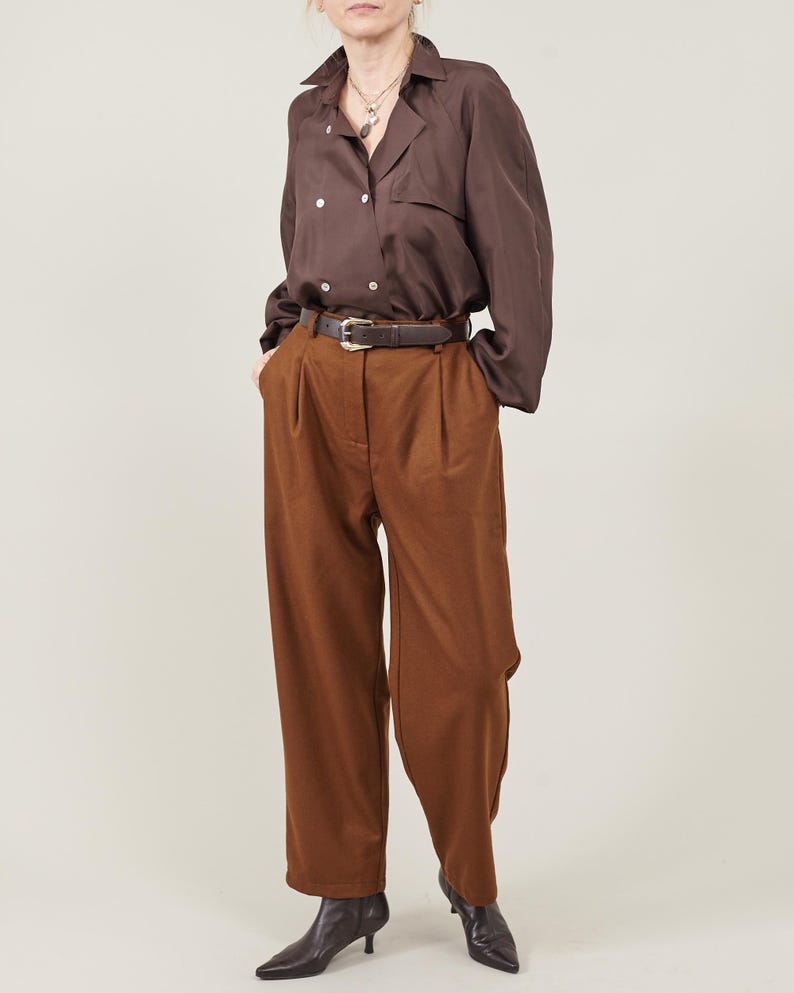May include: A brown button-down shirt with a long sleeve and a brown leather belt. The shirt is tucked into brown wide-leg pants. The person is wearing black heeled boots.