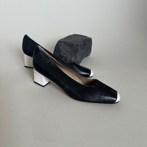 K&ouml;nnte beinhalten: Ein Paar schwarze Lederschuhe mit Absatz und wei&szlig;en Verzierungen an Zehen und Absatz.