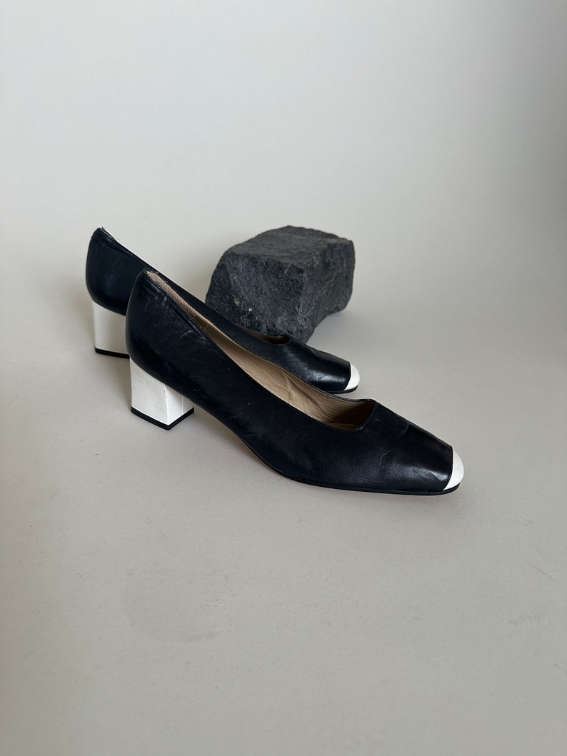 K&ouml;nnte beinhalten: Ein Paar schwarze Lederschuhe mit Absatz und wei&szlig;en Verzierungen an Zehen und Absatz.