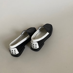 May include: A pair of black and white leather loafers. The shoes have a classic design with a rounded toe, a slip-on style, and a white trim. The heels are adorned with black studs.