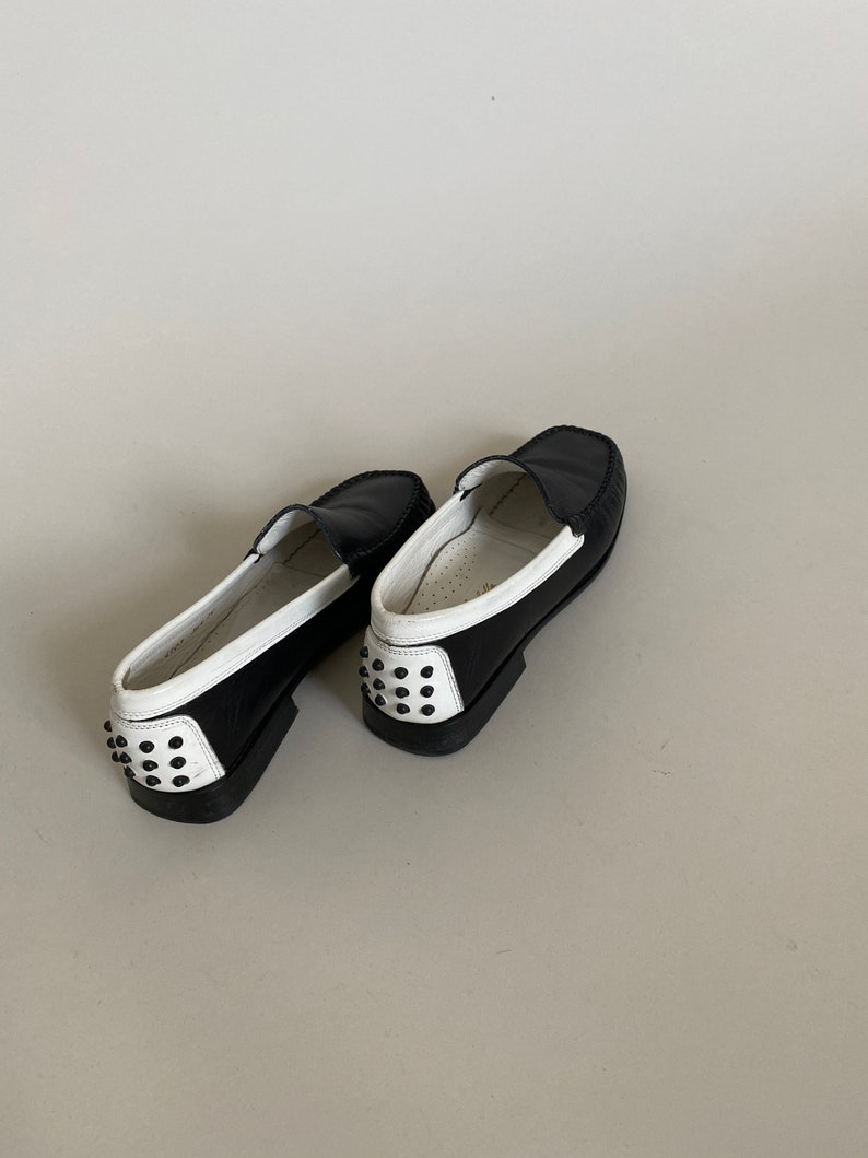 May include: A pair of black and white leather loafers. The shoes have a classic design with a rounded toe, a slip-on style, and a white trim. The heels are adorned with black studs.