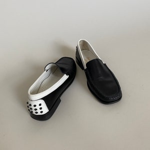May include: Pair of black leather loafers with white trim and a studded heel. The shoes have a classic design with a rounded toe and a slip-on style. The soles are black, and the interior is white.