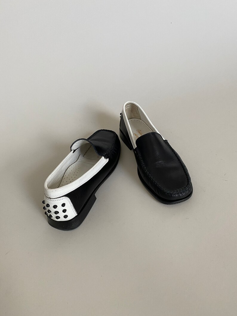 May include: Pair of black leather loafers with white trim and a studded heel. The shoes have a classic design with a rounded toe and a slip-on style. The soles are black, and the interior is white.