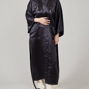 May include: A long black satin robe with embroidered birds and a sash tie. The robe has two pockets and is worn with white trousers.