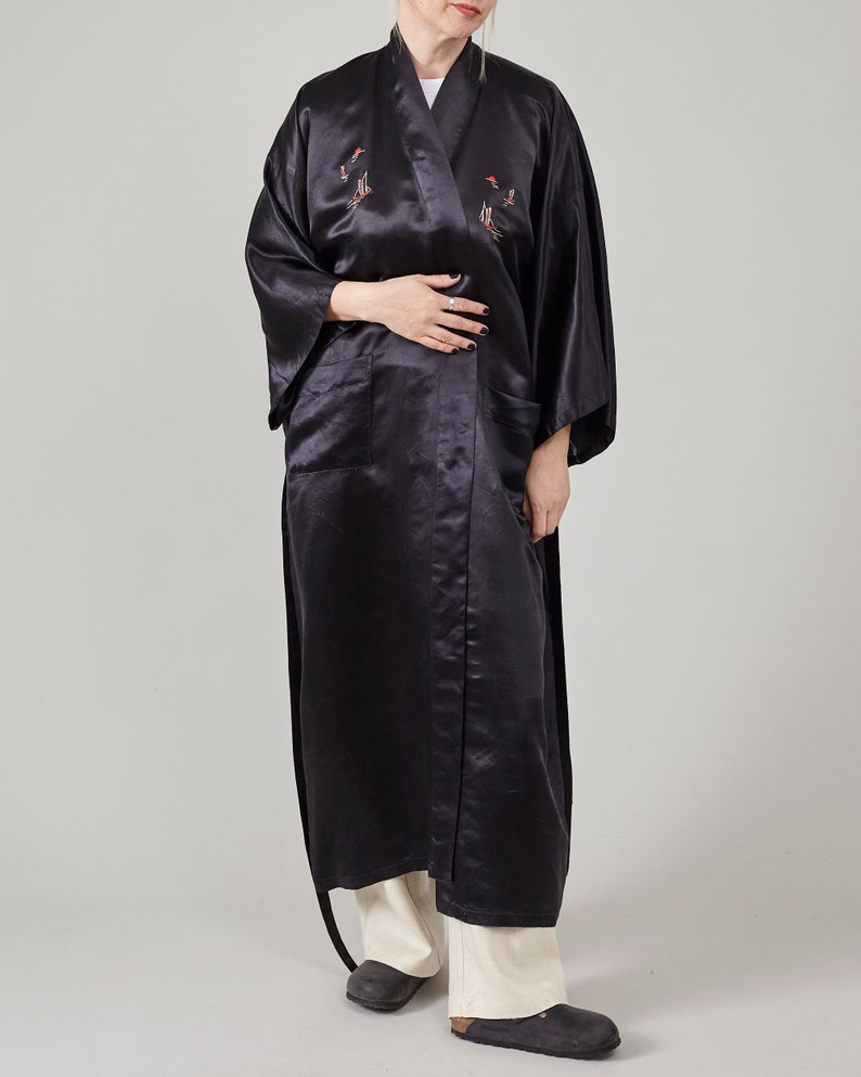 May include: A long black satin robe with embroidered birds and a sash tie. The robe has two pockets and is worn with white trousers.