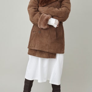 K&ouml;nnte beinhalten: Ein brauner Wildleder- und Lammfellmantel mit einem gro&szlig;en Kragen und einem doppelreihigen Verschluss. Der Mantel wird &uuml;ber einem wei&szlig;en Kleid und braunen Stiefeln getragen.