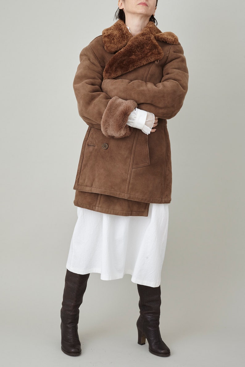 K&ouml;nnte beinhalten: Ein brauner Wildleder- und Lammfellmantel mit einem gro&szlig;en Kragen und einem doppelreihigen Verschluss. Der Mantel wird &uuml;ber einem wei&szlig;en Kleid und braunen Stiefeln getragen.