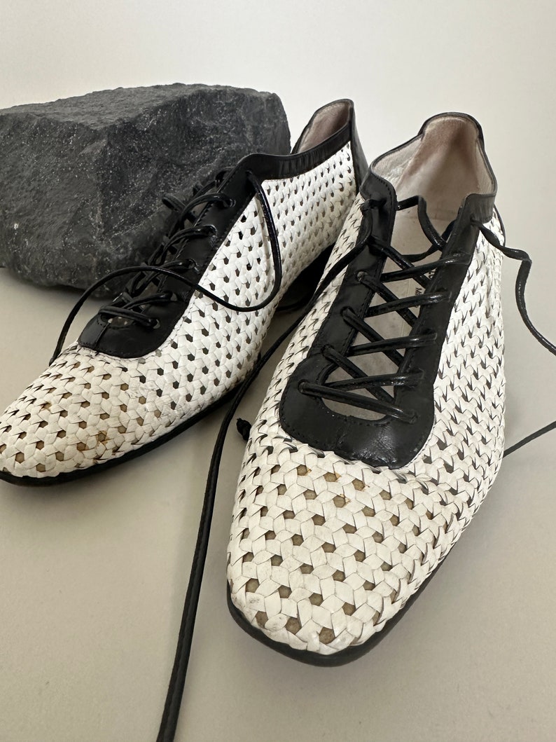 May include: A pair of white and black woven leather shoes with black laces. The shoes have a square toe and a low heel.
