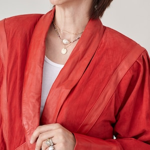 May include: A woman wearing a red leather jacket with a gold chain necklace and a pearl necklace. She is wearing several rings on her fingers.