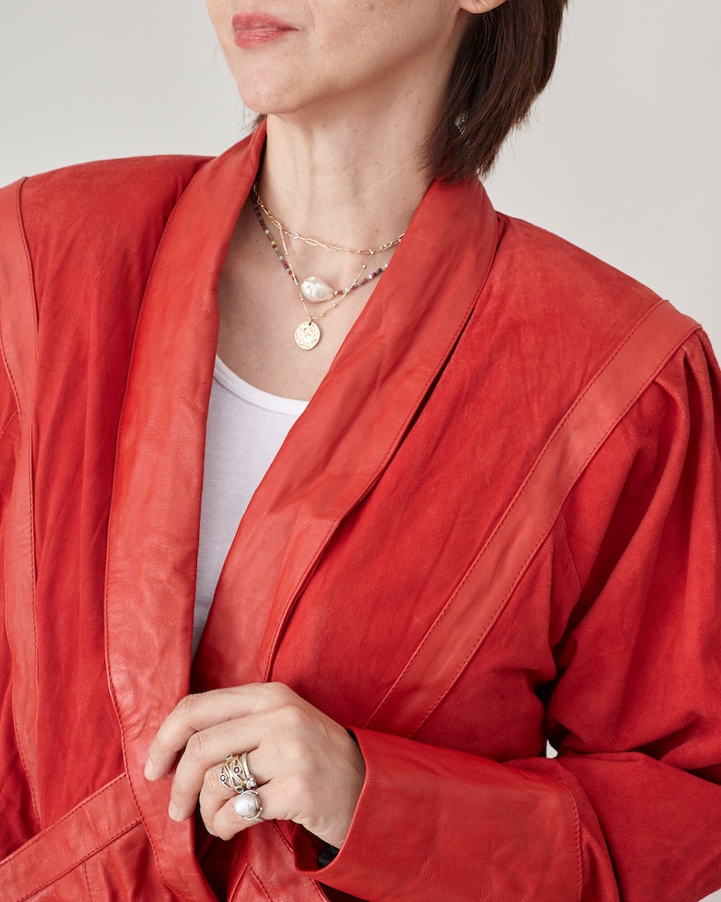 May include: A woman wearing a red leather jacket with a gold chain necklace and a pearl necklace. She is wearing several rings on her fingers.