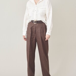 Op de afbeelding: Een wit linnen overhemd met een knoopsluiting en twee borstzakken, gedragen met een bruine linnen broek en een bruine leren riem.