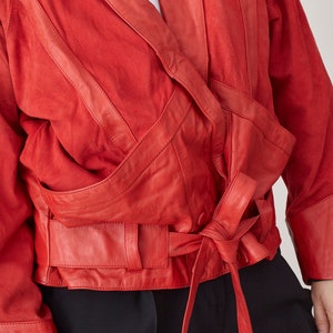 May include: A red leather jacket with a wrap-around design and a tie belt. The jacket has a suede-like texture and features multiple pockets and seams.