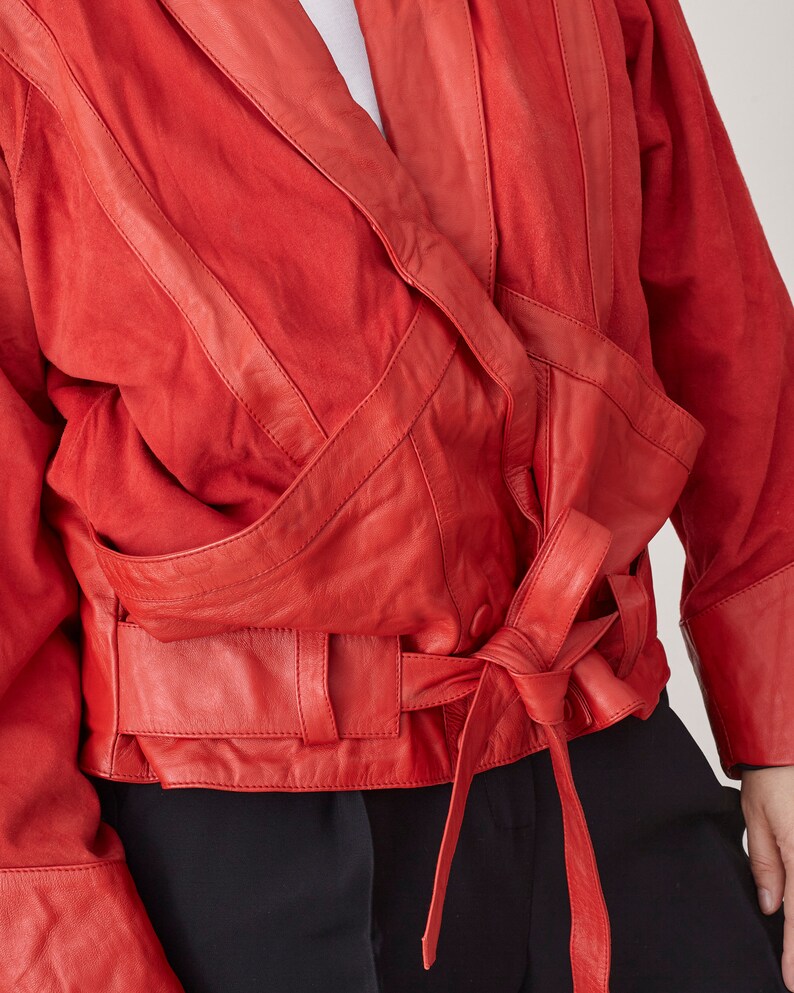May include: A red leather jacket with a wrap-around design and a tie belt. The jacket has a suede-like texture and features multiple pockets and seams.