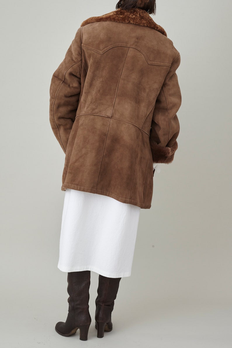 K&ouml;nnte beinhalten: Ein brauner Wildleder-Lammfellmantel mit Pelzkragen und langen &Auml;rmeln. Der Mantel wird &uuml;ber einem wei&szlig;en Kleid und braunen Stiefeln getragen.