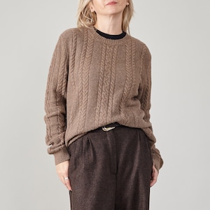 Pode incluir: Um suéter marrom de malha canelada com gola redonda e mangas compridas. O suéter é combinado com calças de pregas marrons escuras e um cinto preto com fivela dourada. Uma camiseta preta também é visível.