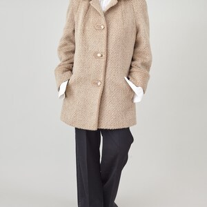 May include: A beige wool coat with a notched collar and four large buttons. The coat has a herringbone pattern and is worn over a white collared shirt and black trousers.