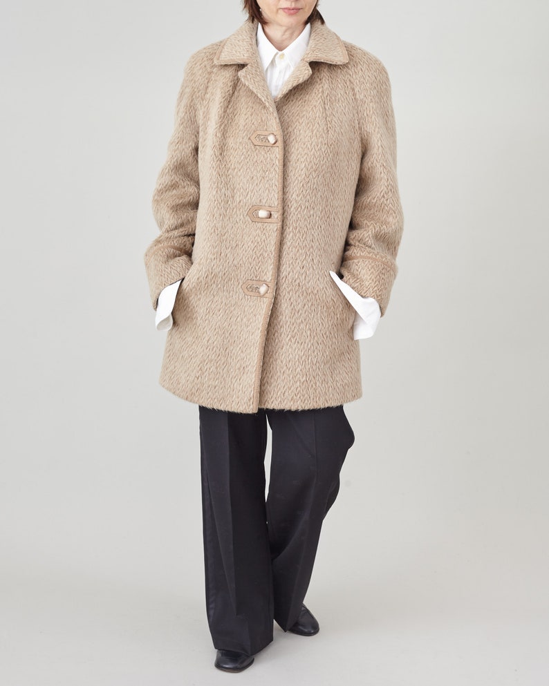 May include: A beige wool coat with a notched collar and four large buttons. The coat has a herringbone pattern and is worn over a white collared shirt and black trousers.