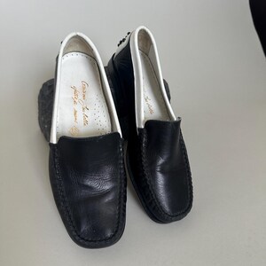 May include: A pair of black leather loafers with white trim around the opening. The shoes have a classic design and appear to be unworn. The interior of one shoe has gold text. The shoes are on a light gray surface.