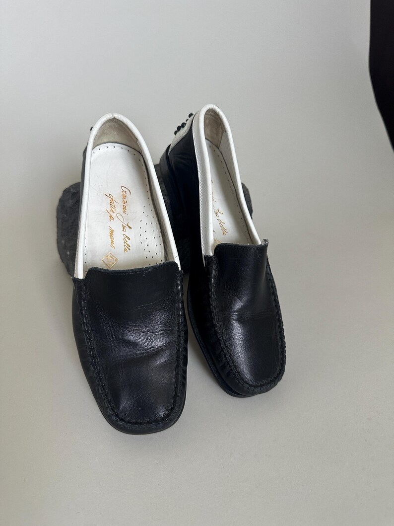 May include: A pair of black leather loafers with white trim around the opening. The shoes have a classic design and appear to be unworn. The interior of one shoe has gold text. The shoes are on a light gray surface.