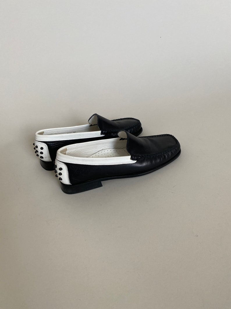 May include: Black and white leather loafers. The shoes have a classic design with a rounded toe and a low heel. The heel features small, round, silver-colored studs. The interior of the shoe is white, and the exterior is black with white accents.