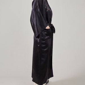 May include: A long black satin robe with a decorative embroidered design on the front. The robe has a sash tie and a pocket on the left side.