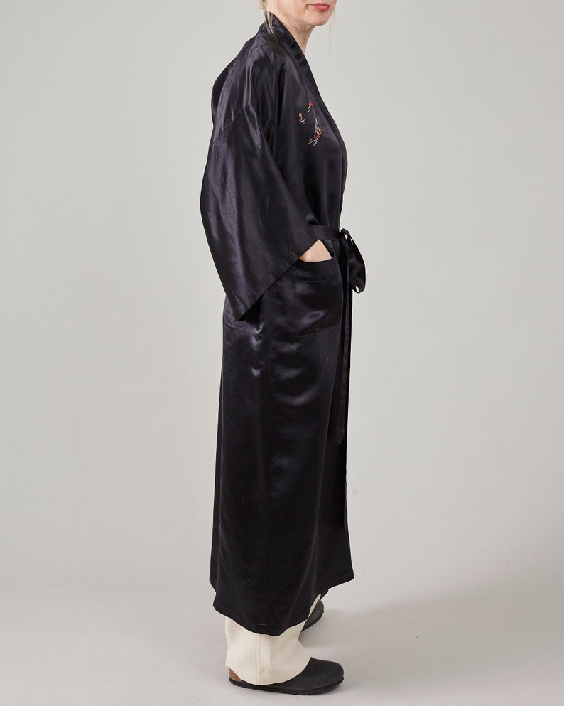 May include: A long black satin robe with a decorative embroidered design on the front. The robe has a sash tie and a pocket on the left side.