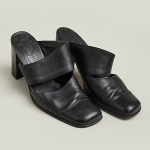 Puede incluir: Par de mules de tac&oacute;n de cuero negro con punta cuadrada y una sola tira en la parte superior. Los zapatos tienen un tac&oacute;n grueso y son de la marca "Betsy Johnson".