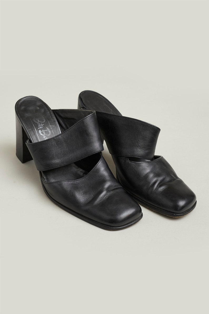Puede incluir: Par de mules de tac&oacute;n de cuero negro con punta cuadrada y una sola tira en la parte superior. Los zapatos tienen un tac&oacute;n grueso y son de la marca "Betsy Johnson".