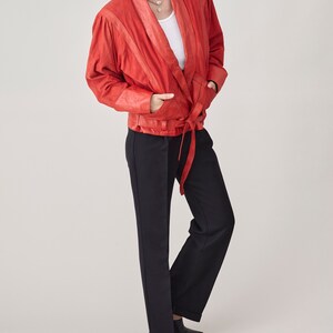 May include: A red leather wrap jacket with a long tie belt. The jacket has a white undershirt and black pants.