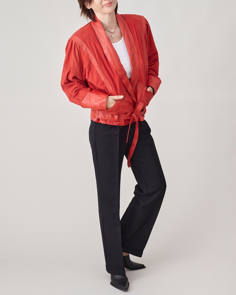 May include: A red leather wrap jacket with a long tie belt. The jacket has a white undershirt and black pants.