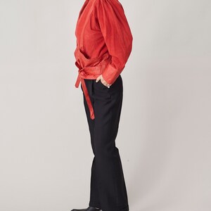 May include: A red suede jacket with a wrap-style front and a self-tie belt. The jacket has long sleeves and a relaxed fit. The model is wearing black pants.