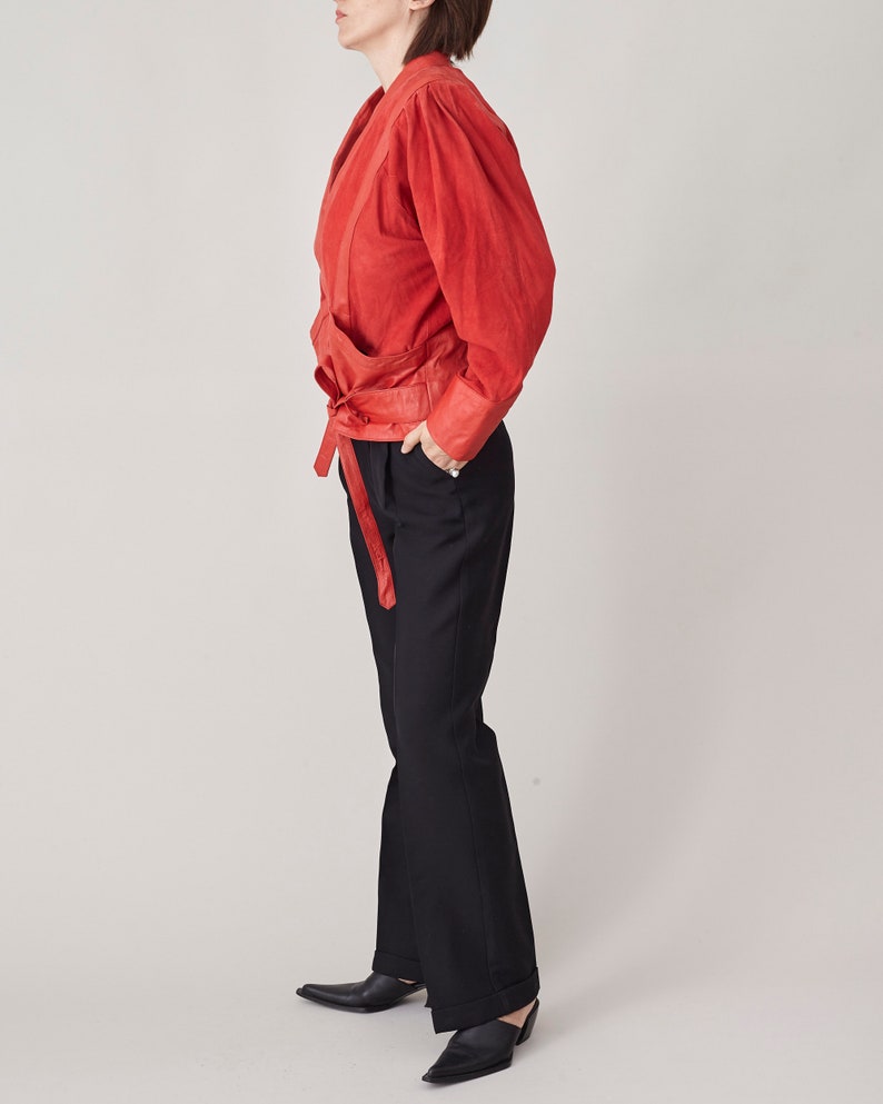 May include: A red suede jacket with a wrap-style front and a self-tie belt. The jacket has long sleeves and a relaxed fit. The model is wearing black pants.