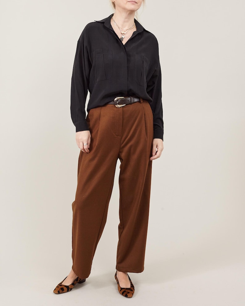 May include: A woman wearing a black button-down shirt with a brown belt and brown wide-leg pants. She is wearing brown and black leopard print flats.