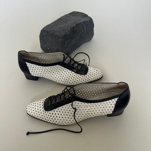 May include: A pair of white and black lace-up shoes with a perforated design. The shoes have a low heel and a rounded toe.