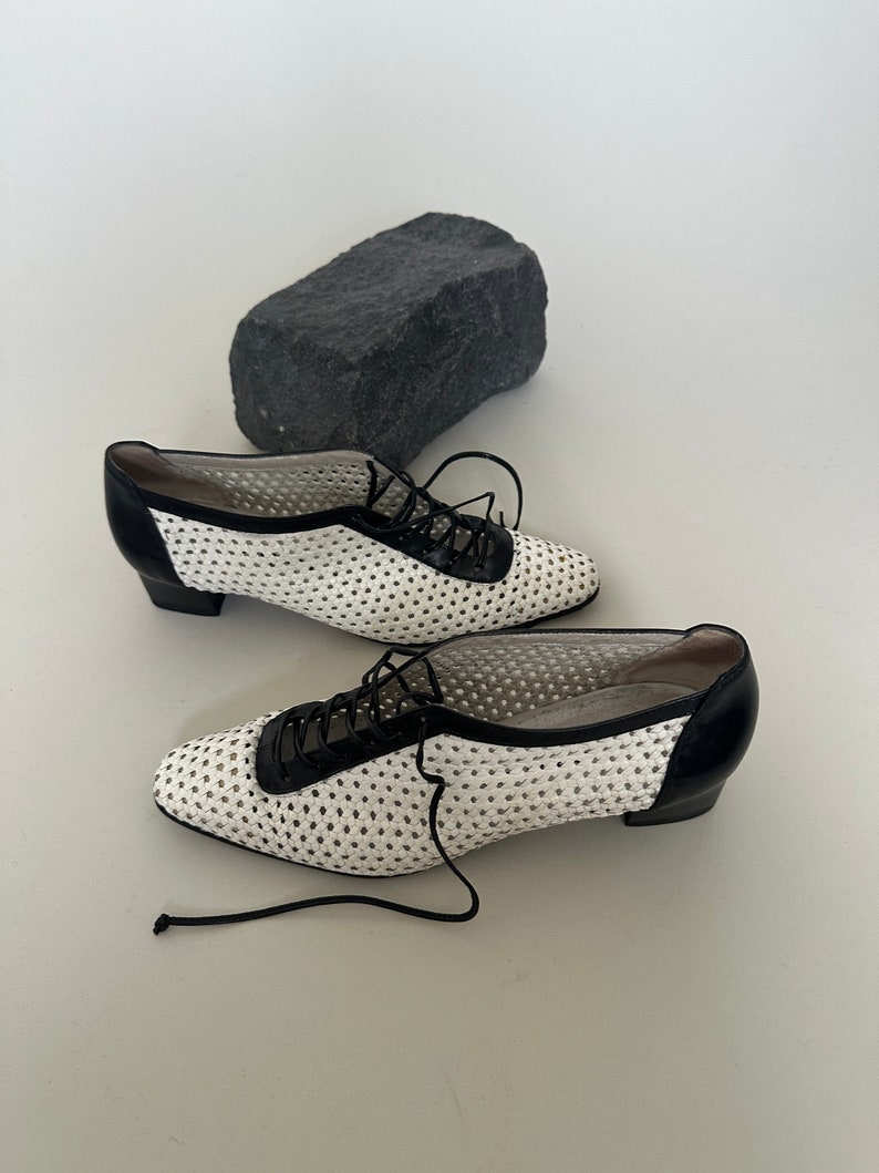 May include: A pair of white and black lace-up shoes with a perforated design. The shoes have a low heel and a rounded toe.