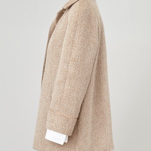 May include: A beige wool coat with brown trim around the cuffs and collar. The coat has a relaxed fit and a slightly oversized silhouette.