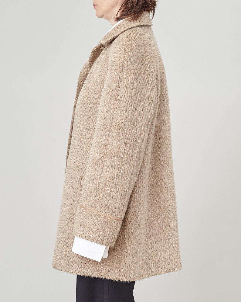 May include: A beige wool coat with brown trim around the cuffs and collar. The coat has a relaxed fit and a slightly oversized silhouette.